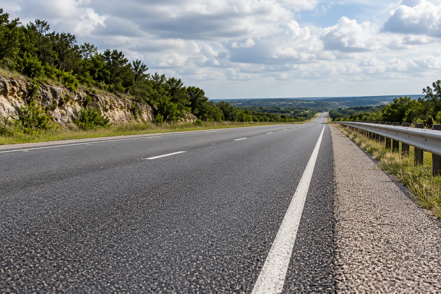 An empty paved roadway with a dashed white lane divider, solid shoulder line and metal guardrail.